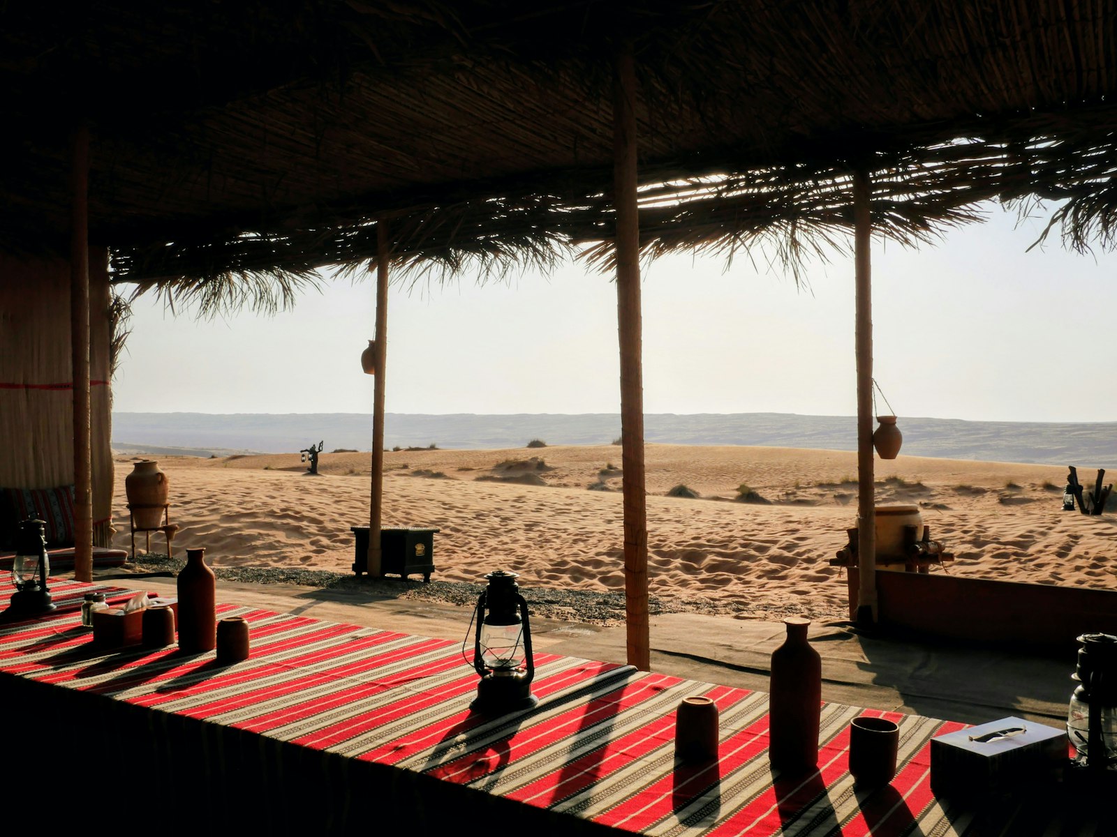 Bedouin desert camp interior with woven red carpets, lanterns and palm-thatch shade looking out over Oman's golden sand dunes